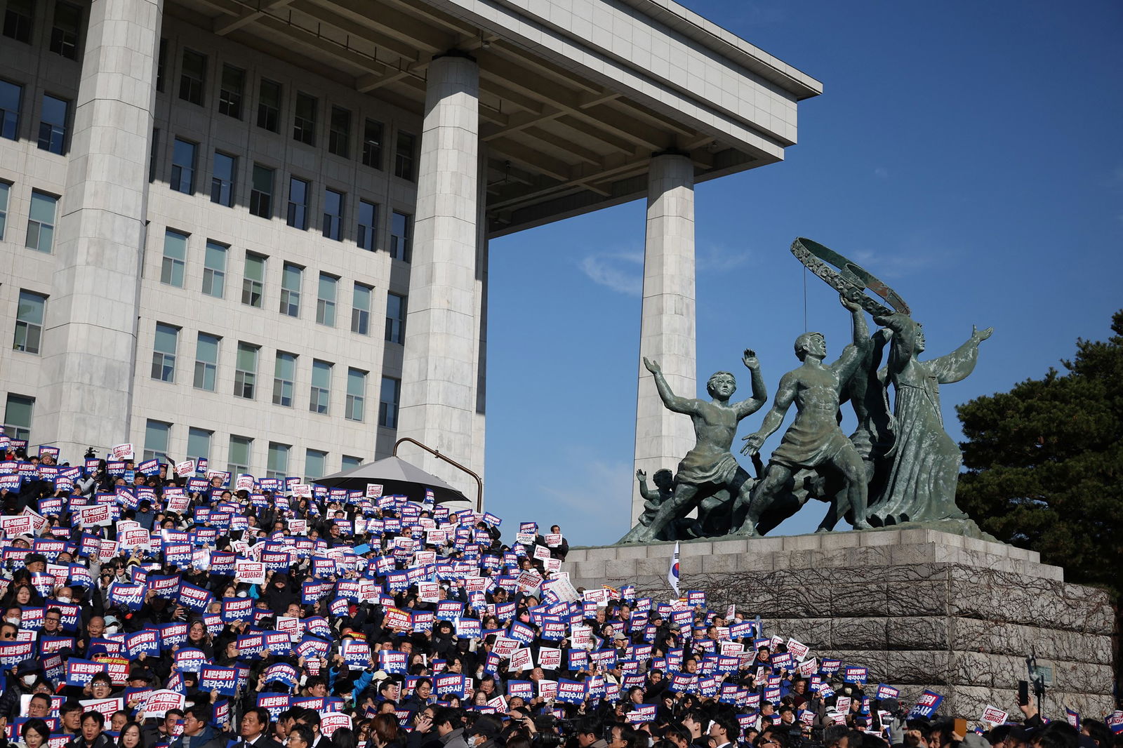 the rally shown on the steps of a tall building with a green metal statue of people with their arms up