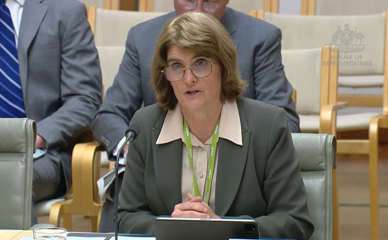 A woman with a short light brown bob and grey suit sits at a desk with a small microphone in front of her.