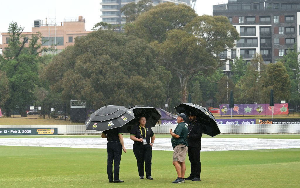 Match officials stand under umbrellas in the rain at a cricket field.