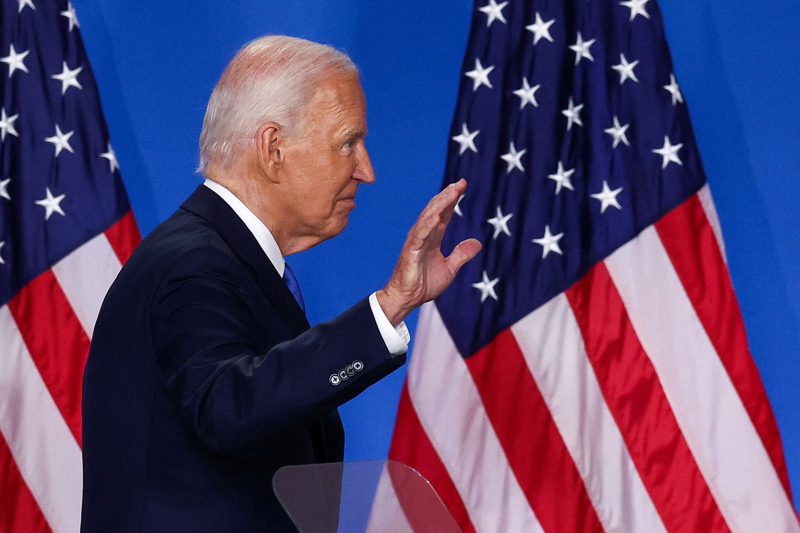 A man turned to the side with his hand out, walking in front of two USA flags.