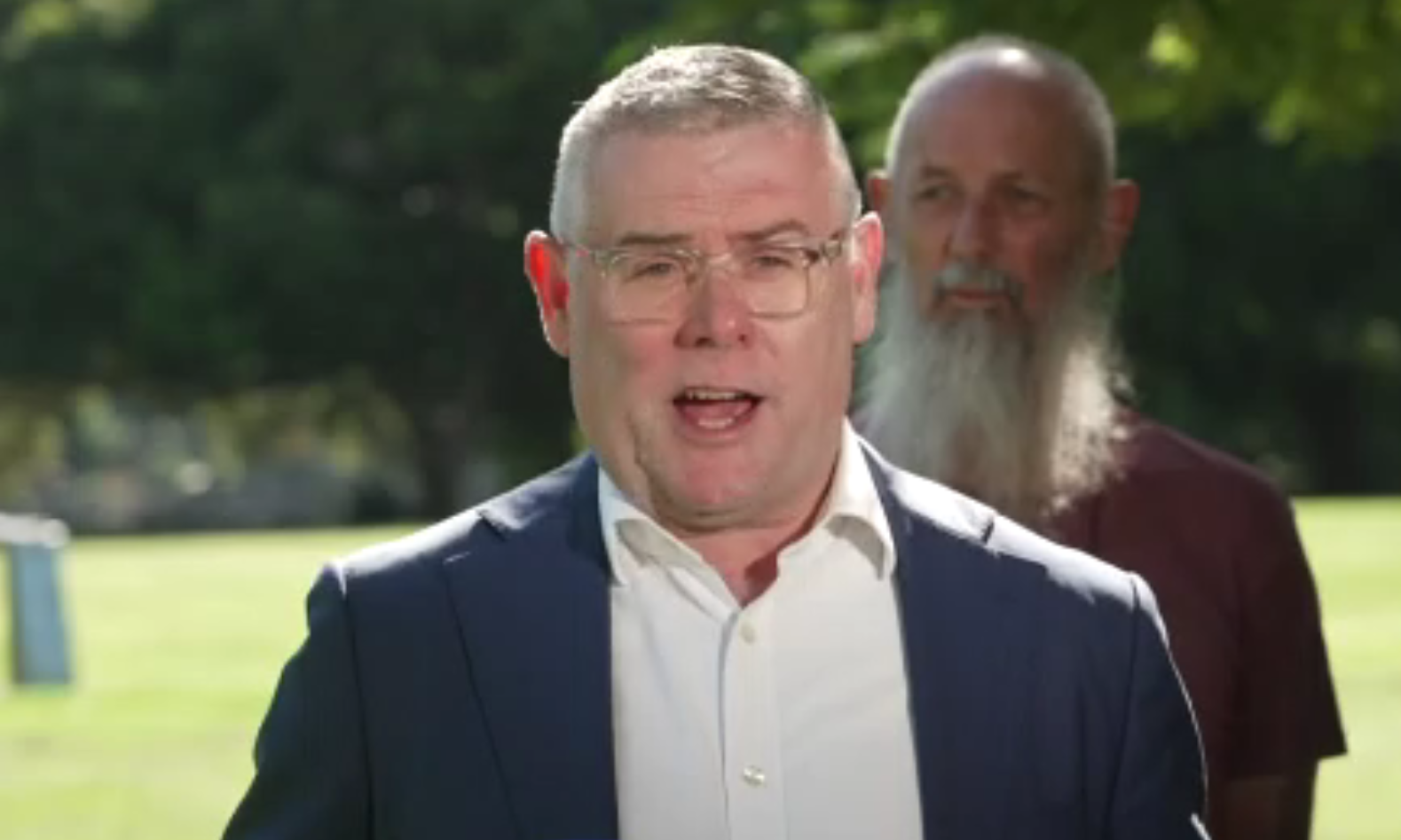 Murray Watt speaks in a park in front of a bearded man.