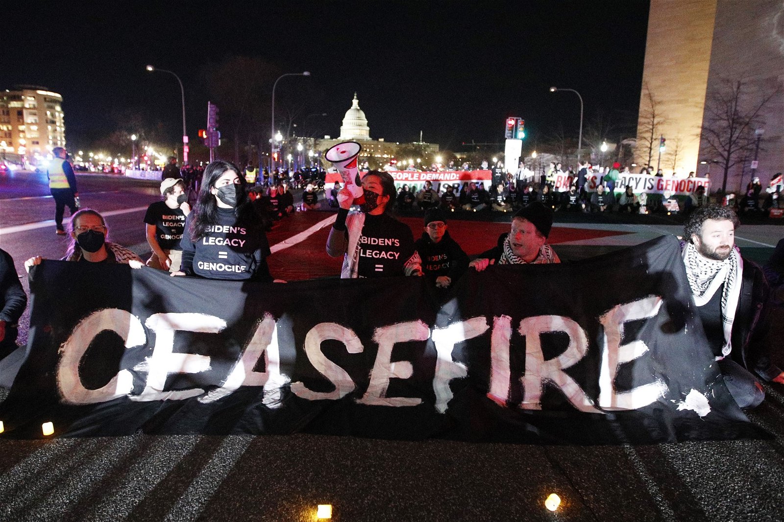 Protesters wearing black T shirts hold a sign with CEASFIRE on a road at night.