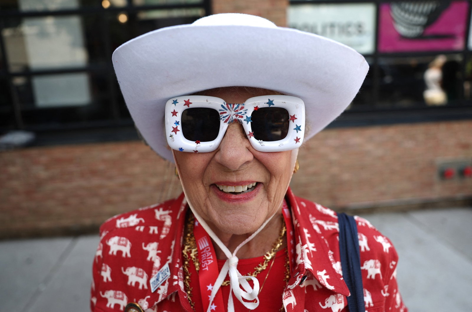 A woman wears a large white hat and novelty sunglasses with blue and red stars.