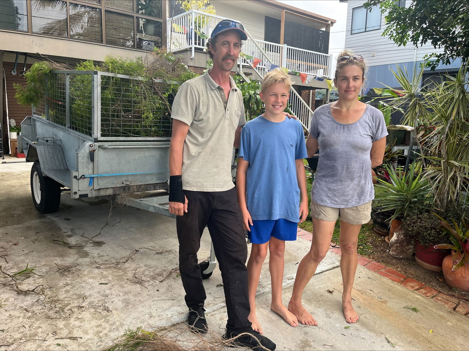 A man and a woman standing on their driveway with their son. Behind them is a trailer full of green waste. 