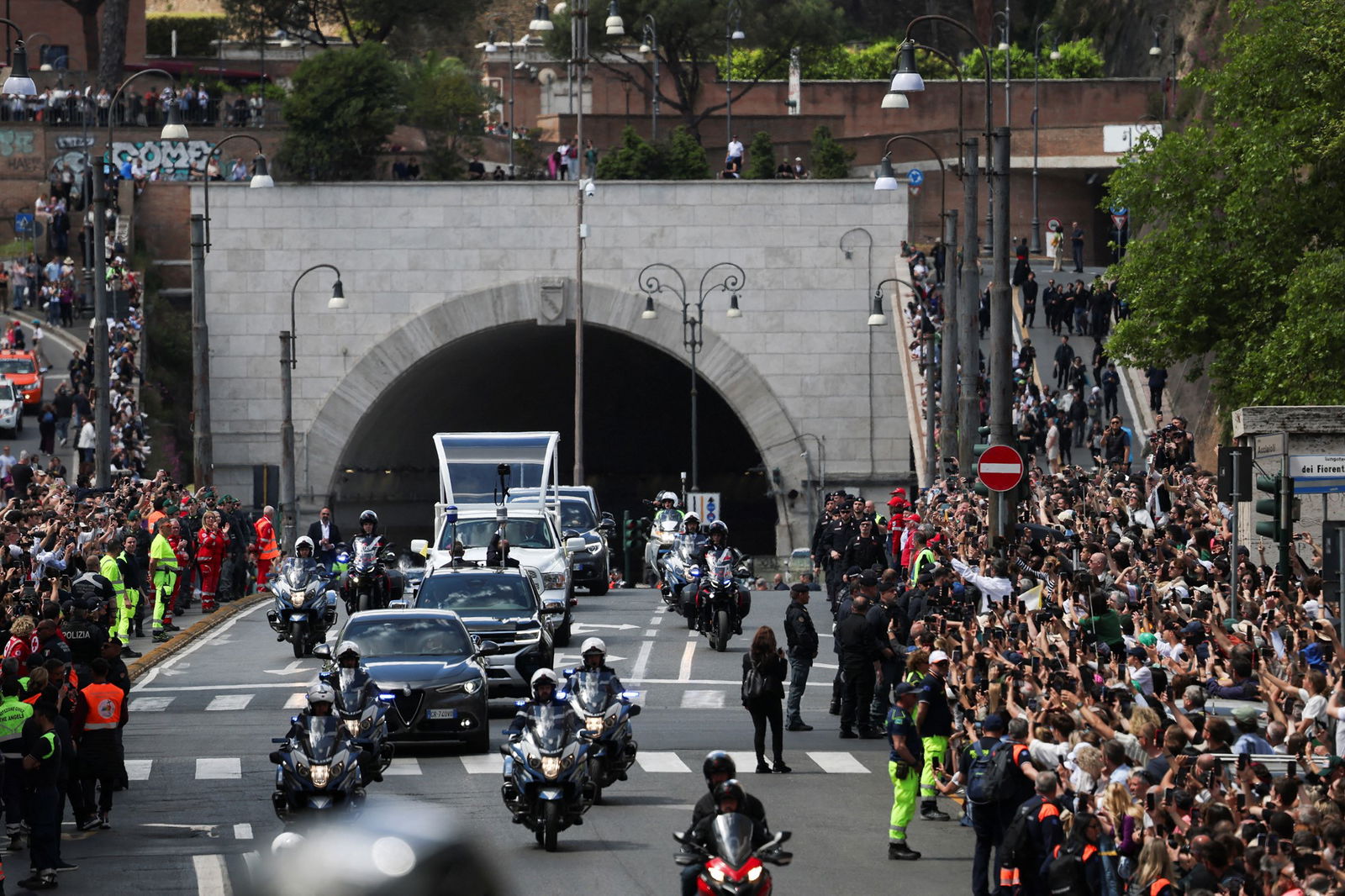 Crowds watch a hearse and police vehicles on a road.