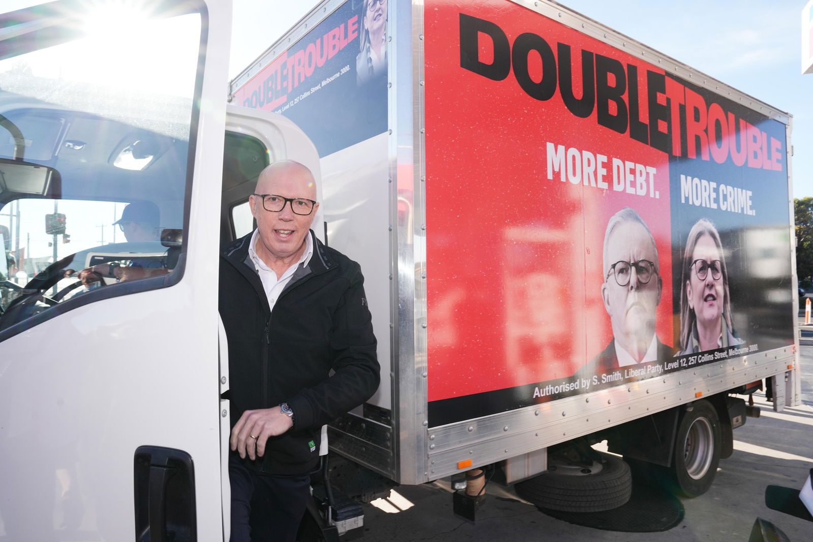Peter Dutton smiling while standing in the doorway of a truck with a trailer adorned with images of Anthony Albanese and Jacinta Allen and the words 'double trouble'