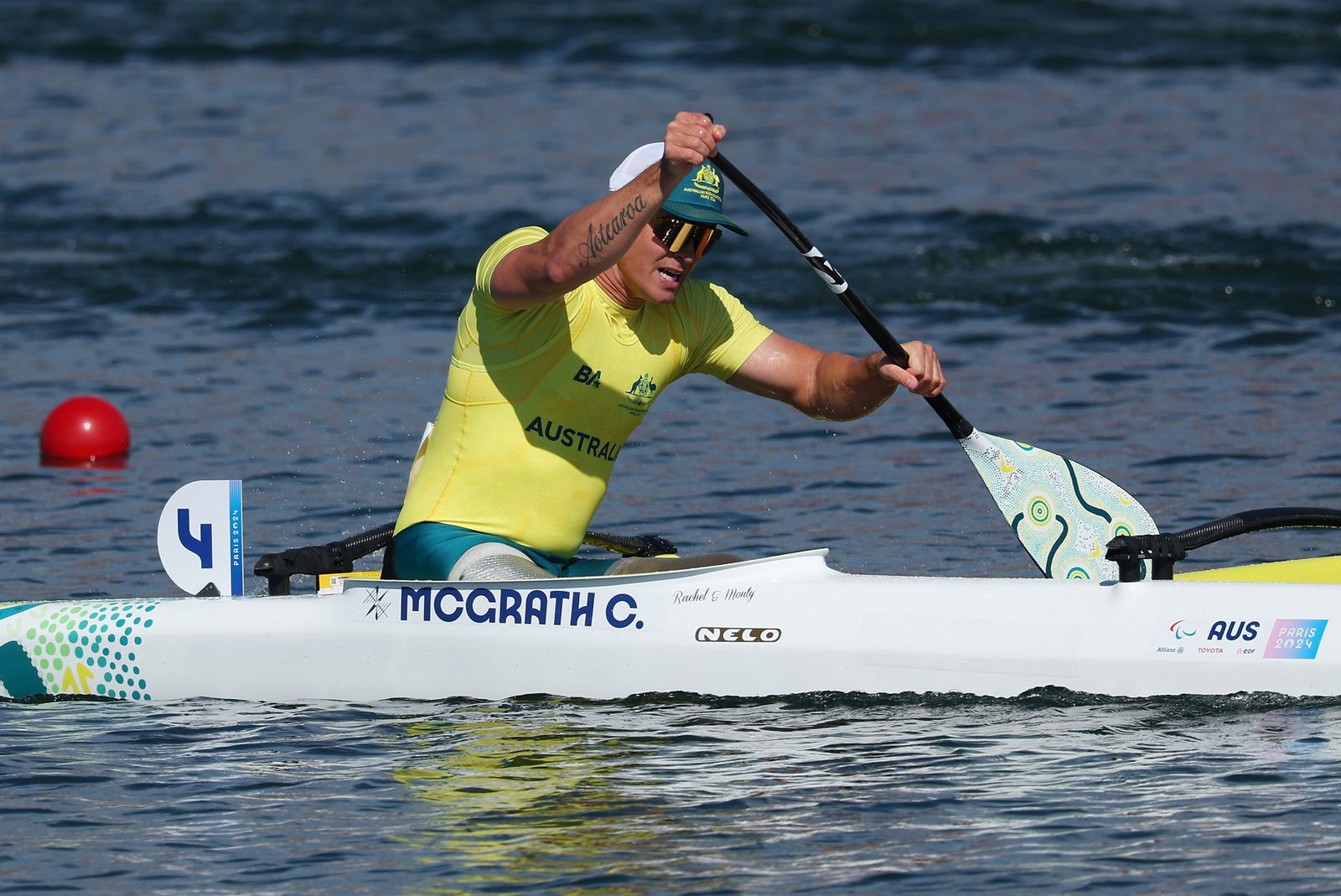 A man is padding in a kayak on rippling grey water. He is wearing a green cap, reflective sunglasses, a yellow tightly fitting sports top and green pants. He has both hands on an oar and is plunging it into the water to his left. The oar has a black handle and a white paddle.