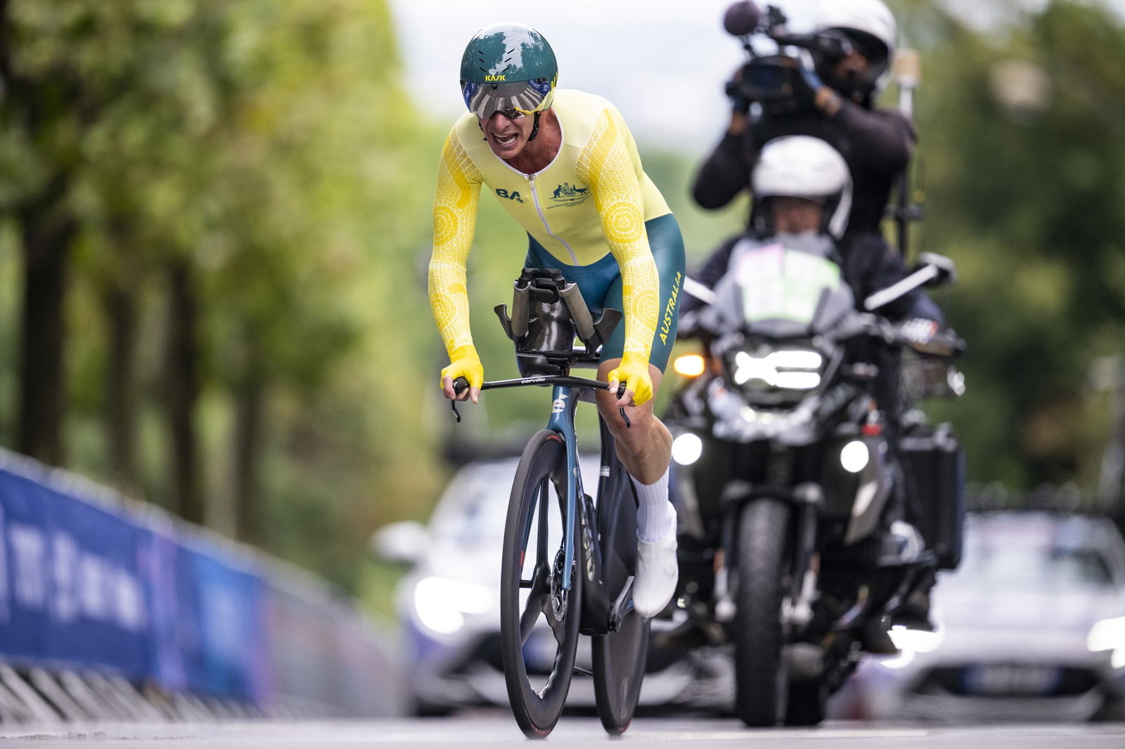 A man with a green helmet, yellow shirt and green pants rides a racing bike. He is wearing sunglasses with a grimaced expression and has one leg. Behind him, there is a motorcycle with a rider and a man standing behind him with a camera, videoing the cyclist. There are cars behind the motorcycle and trees in the background. The road is bordered by portable fences.