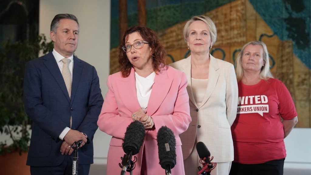 Amanda Rishworth speaking in pink blazer with Mark Bulter, Tanya Plibersek standing beside her, and a blonde home care worker in red union shirt.