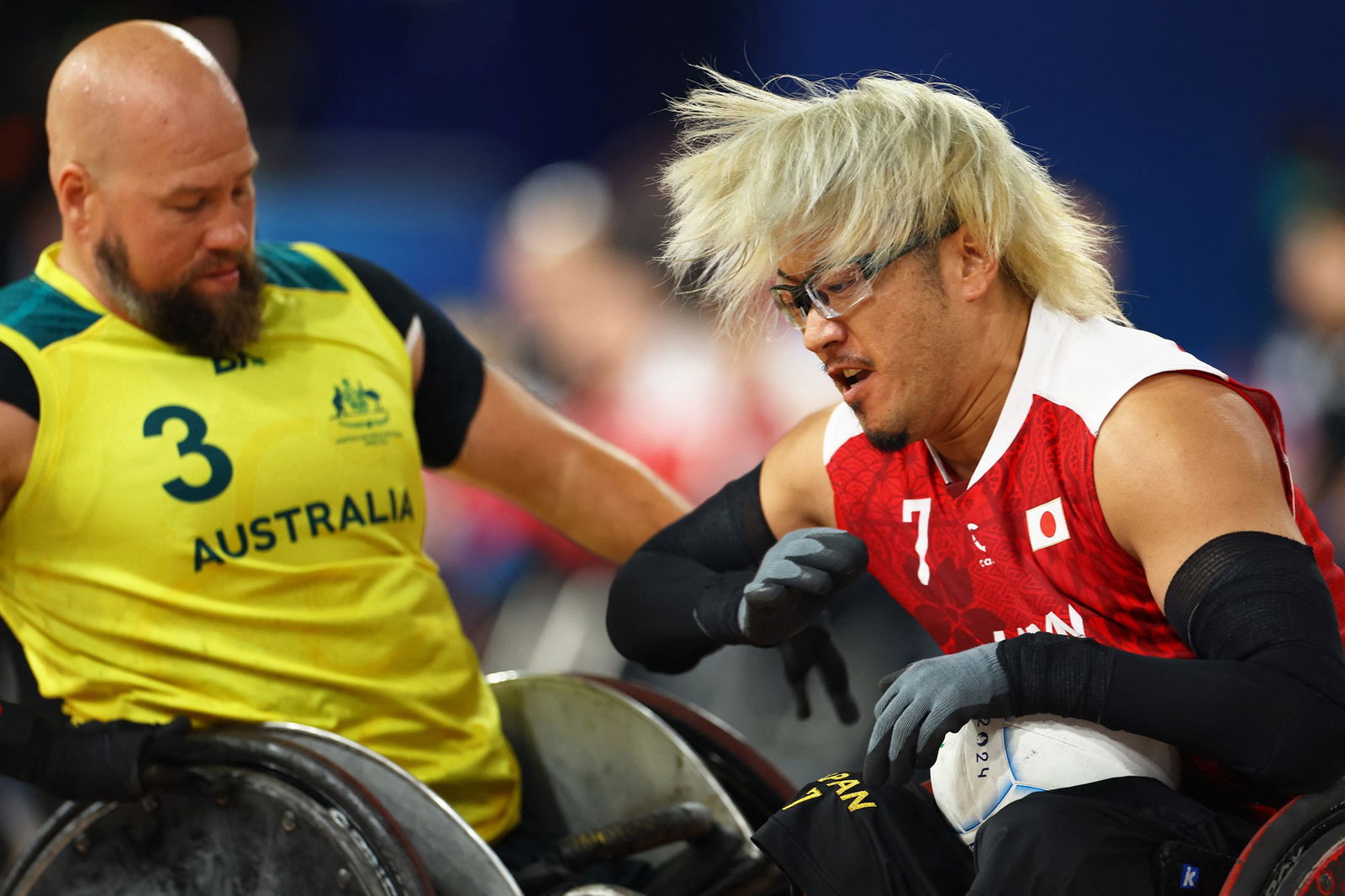 A Japanese male wheelchair rugby player holds the ball while an Australian defender hits him with his chair.