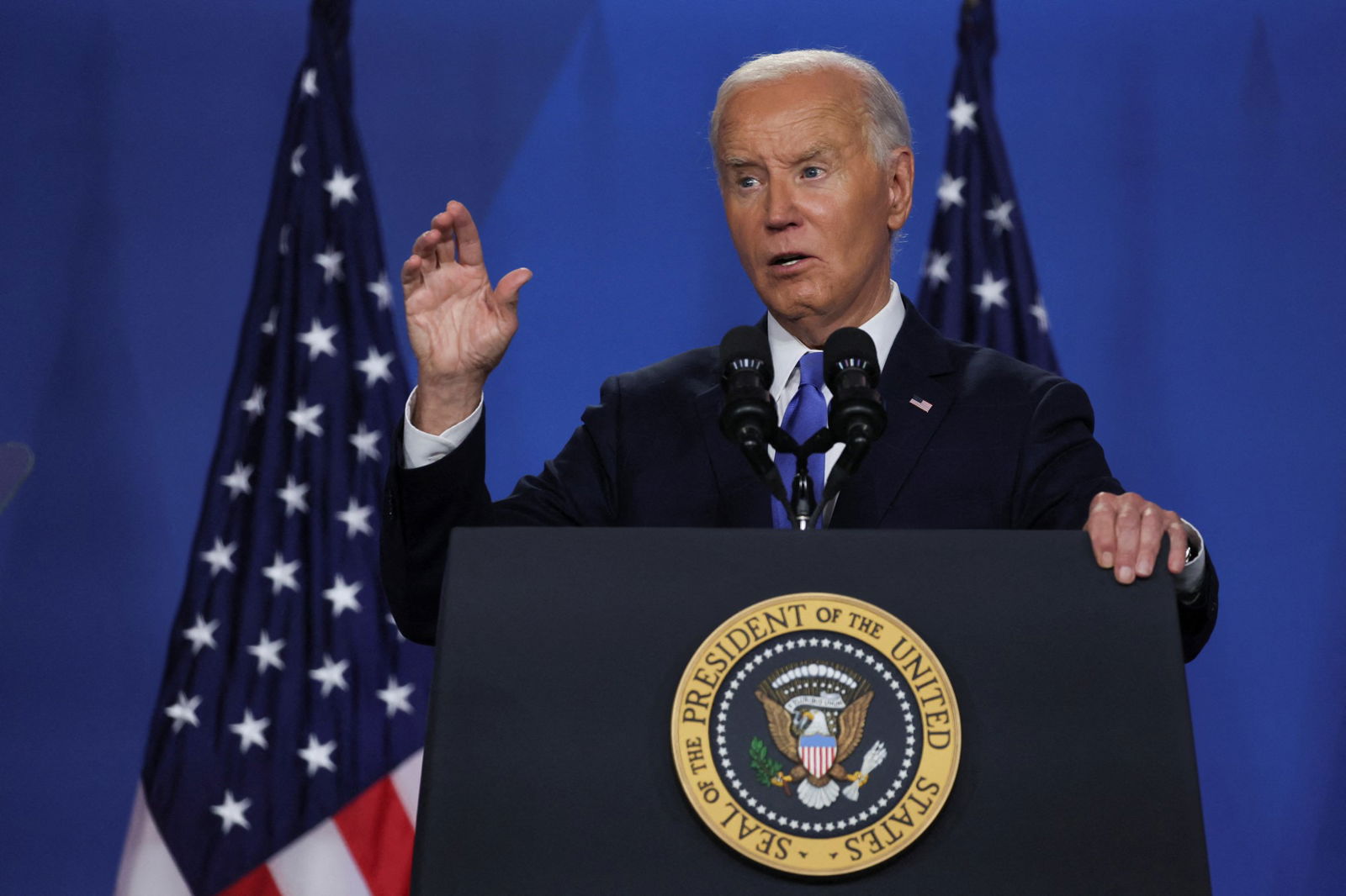 A man in front of a podium with two USA flags behind it.