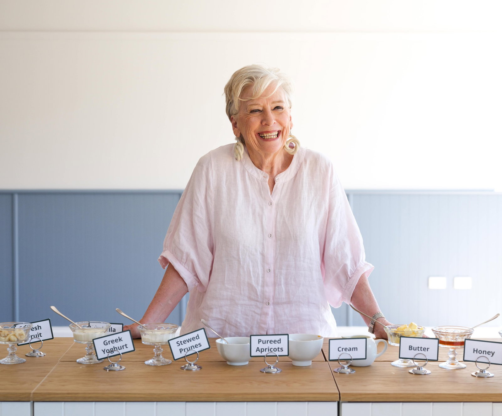 A smiling Maggie Beer stands at a bench before an assortment of bowls of food, including Greek yoghurt and pureed apricots. 