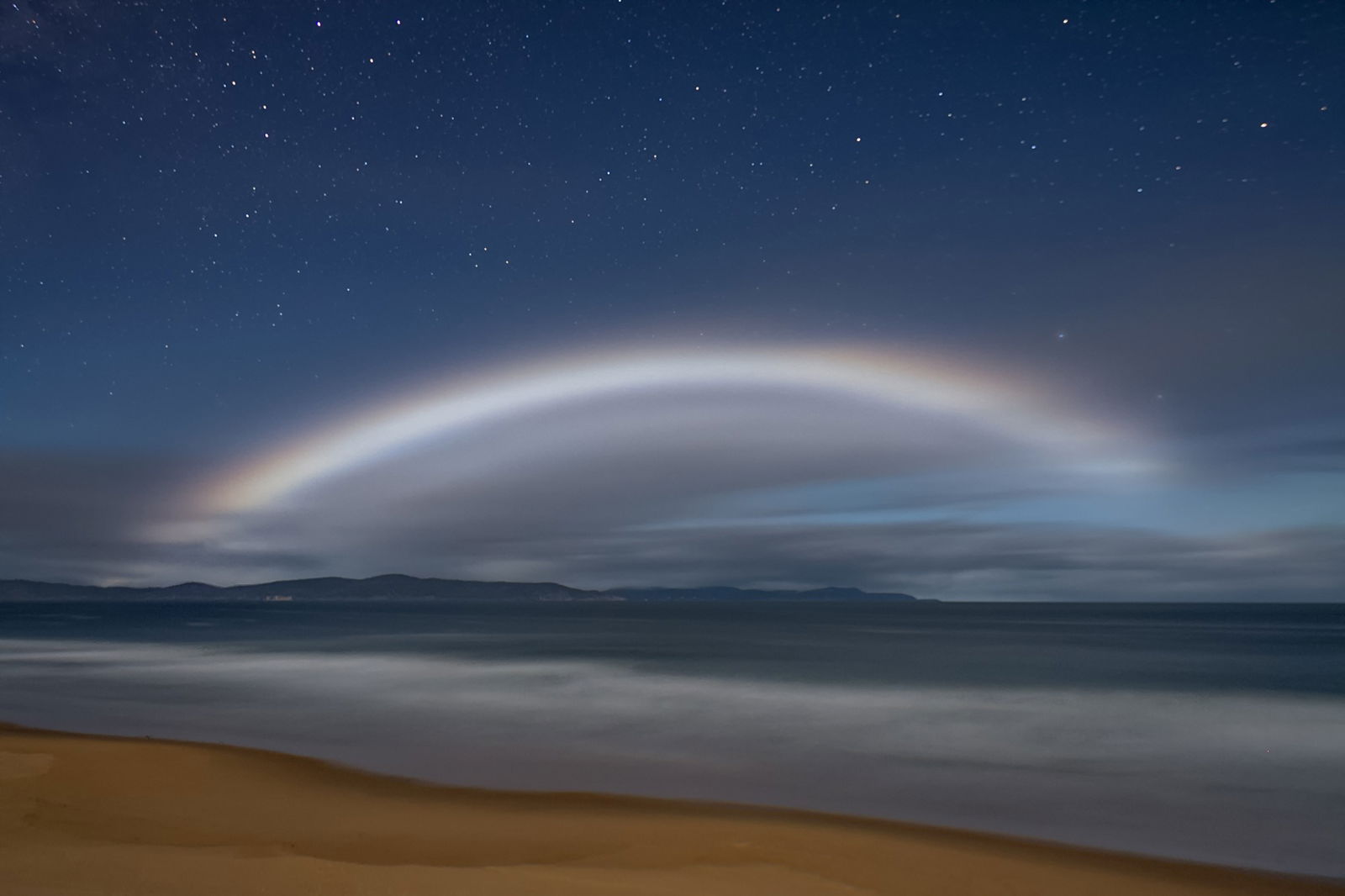 A silver arc of light seen in the evening sky over a beach