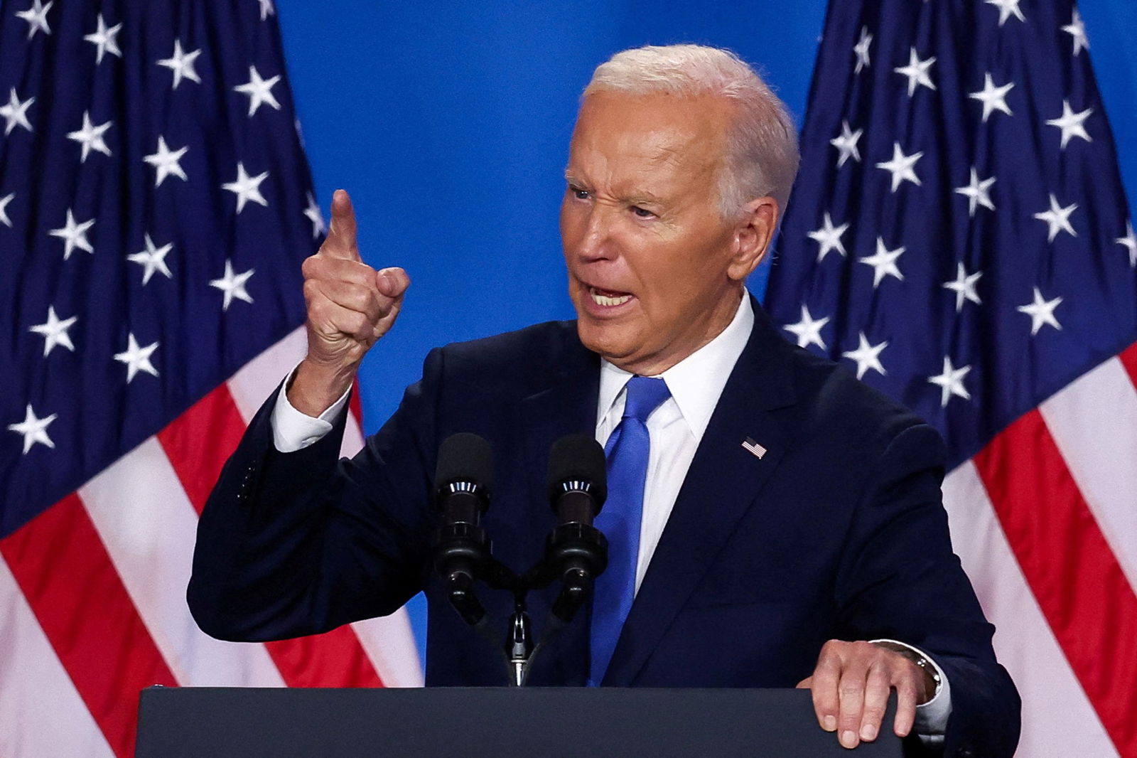 A man in a blue suit with two USA flags behind him