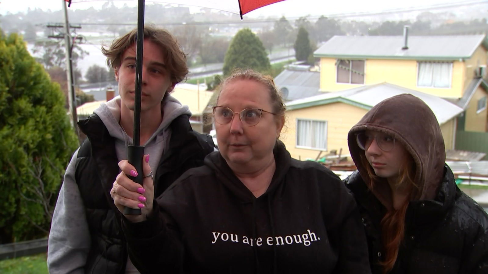 A woman holds an umbrella and speaks outside a home.