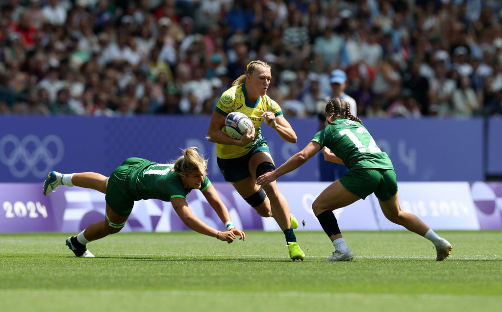 A blonde woman in a gold shirt splits two defenders in green shirts. 