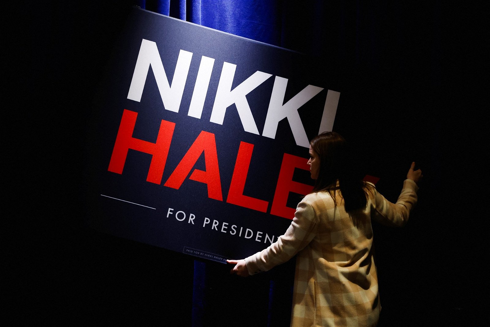 A woman holds up a 'Nikki Haley for President' sign in a shadowy room.