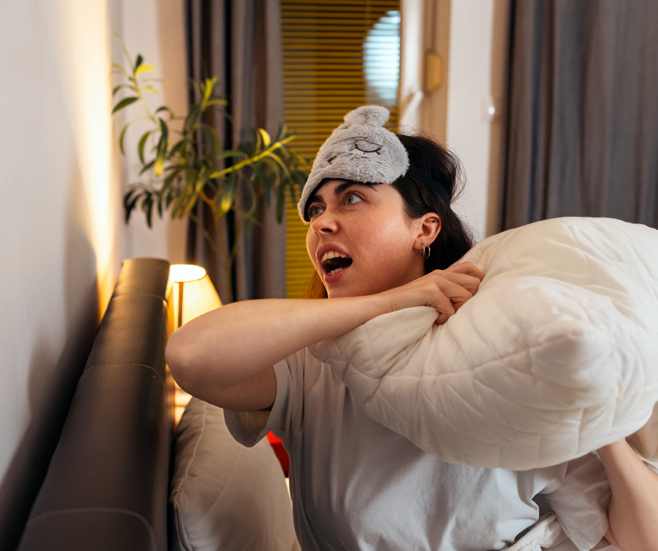 A woman wearing a sleeping mask looks angry in bed and makes to hurl a pillow at a wall.