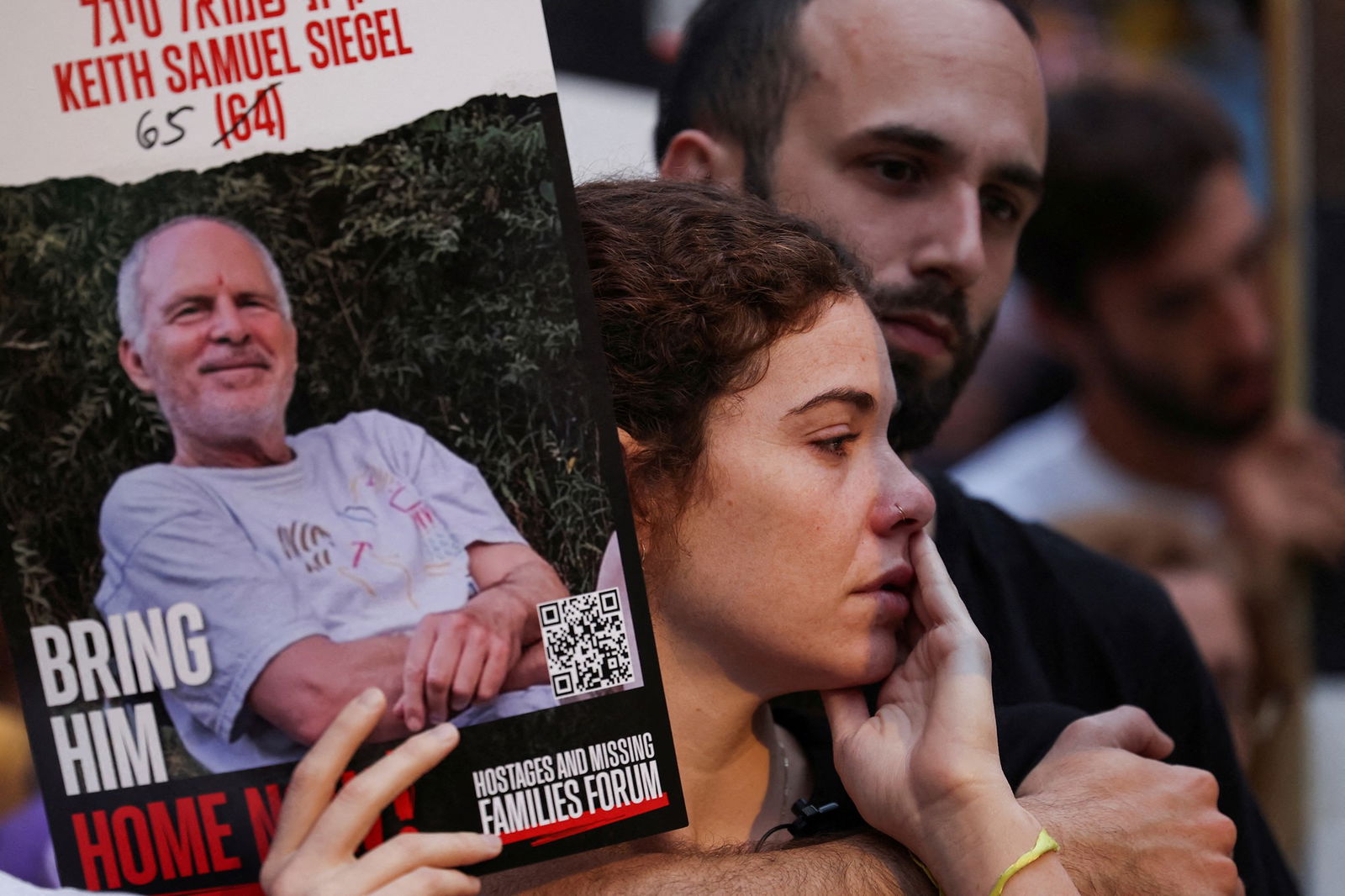 A man and woman hugging, with the woman holding a poster showing another man's face