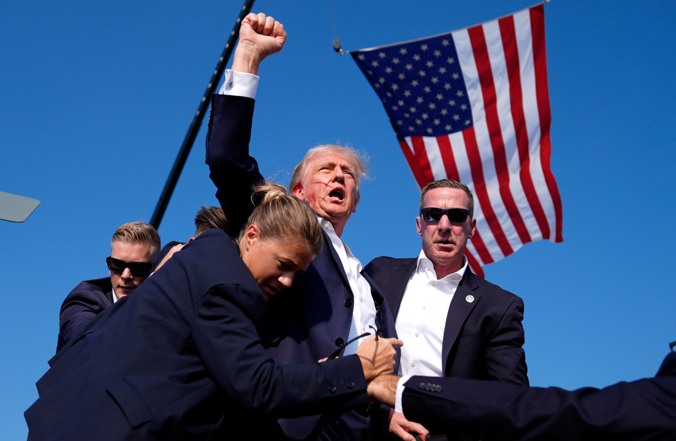 President Trump raises a fist in the air as he bleeds from the ear and Secret Service agents rush him from a stage.