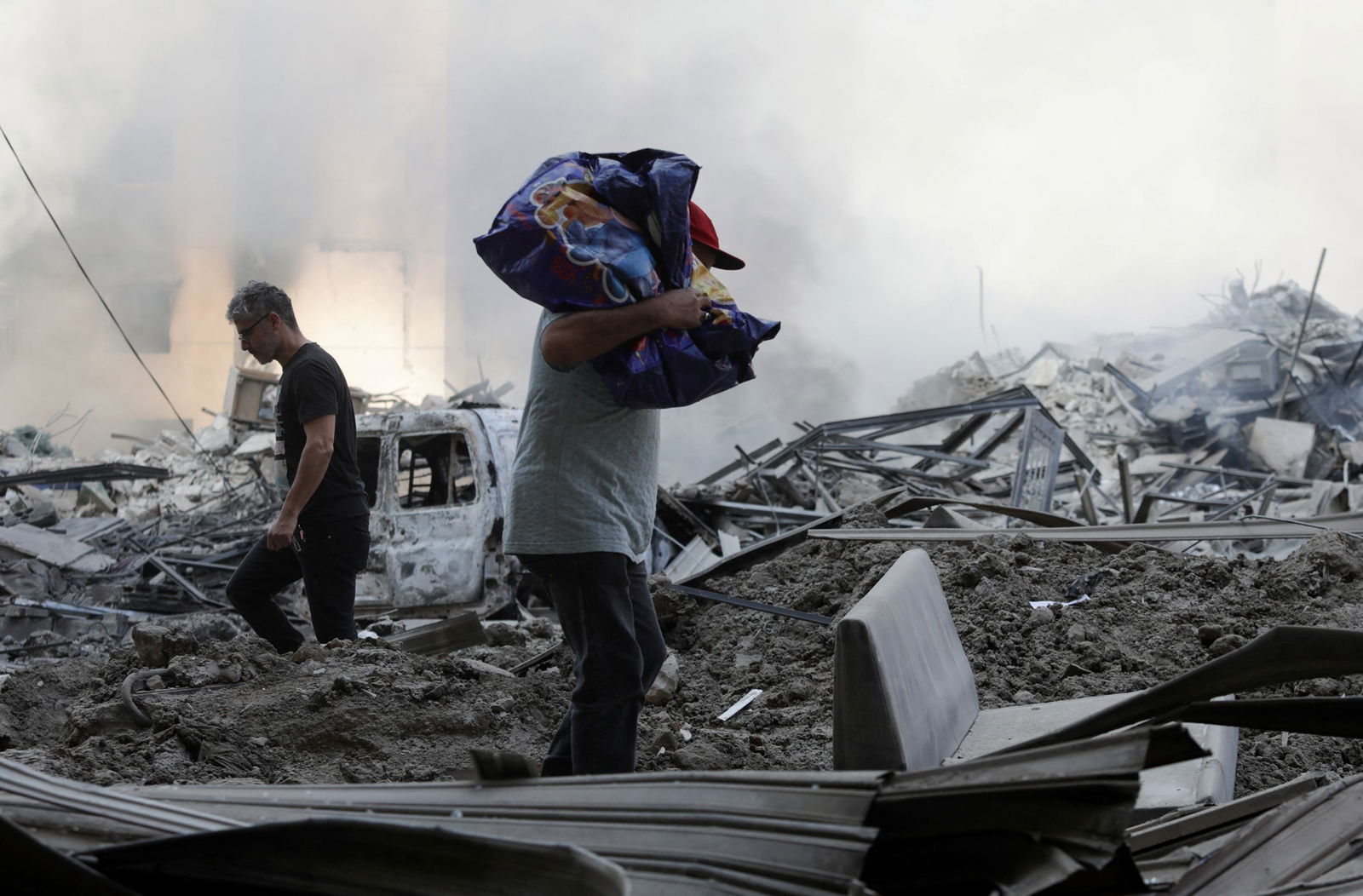 Two men walk amongst smoking rubble, one carrying a bundle