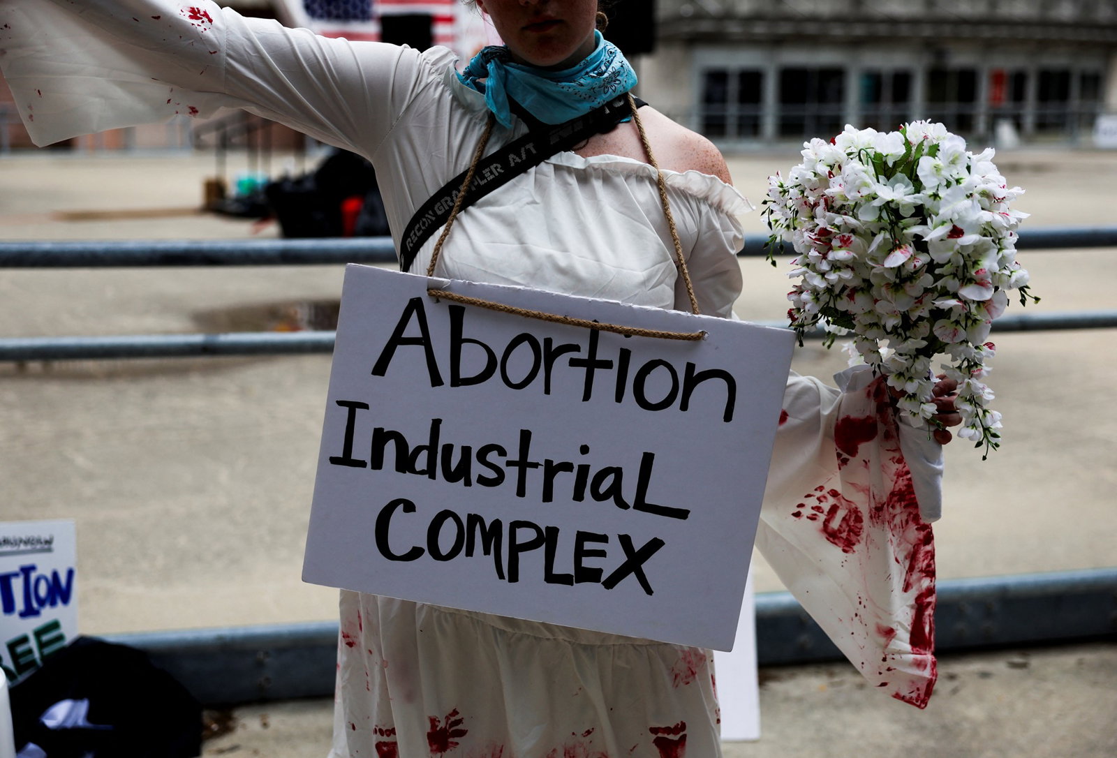 A protester wearing a white dress with red streaks holds a bouqet of flowers. A sign reading 'Abortion industrial complex' hangs around her neck