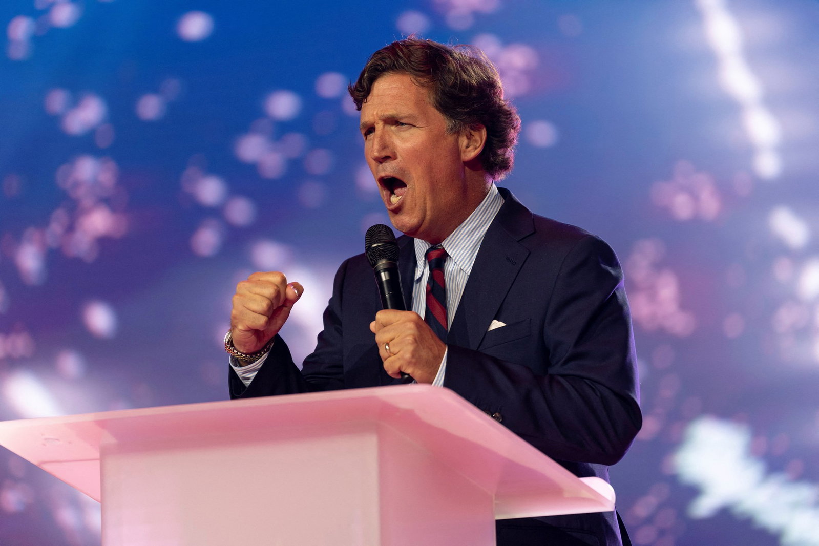 Tucker Carlson speaks emphatically into a handheld microphone, clenching one fist.