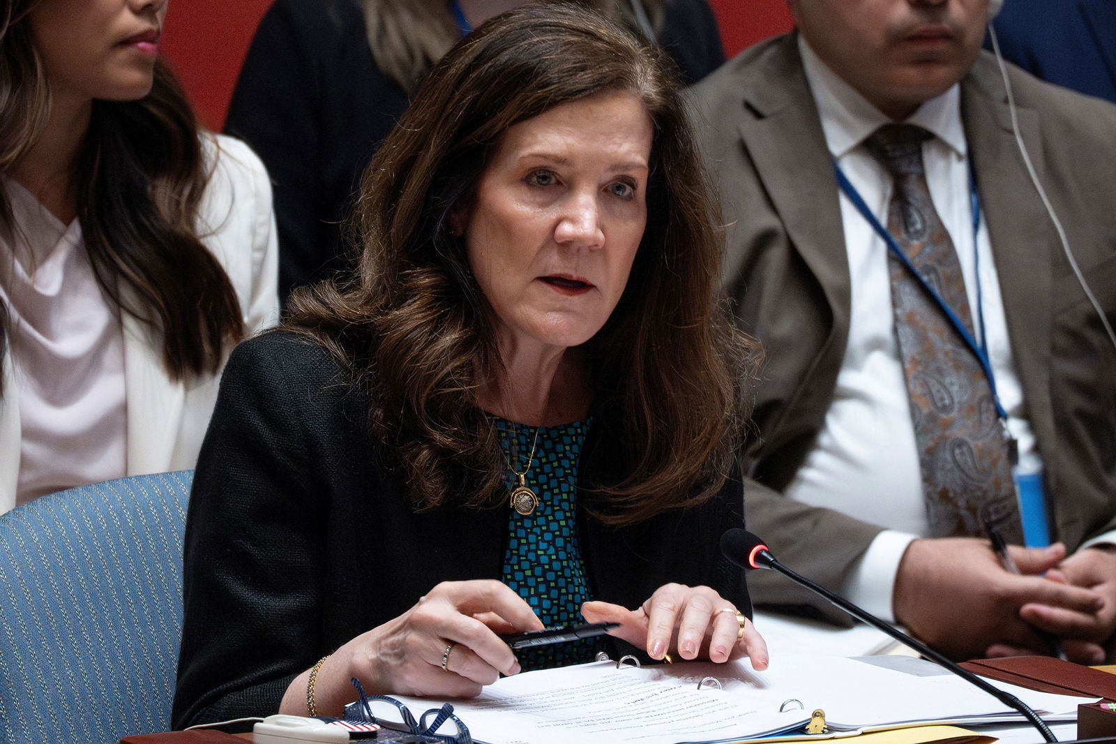 A woman with long, brunette hair rests her hands on an open binder filled with pages as she speaks at a desk