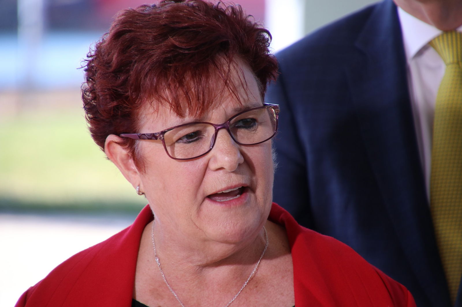 A woman with dyed red hair and glasses stands at podium addressing the press.