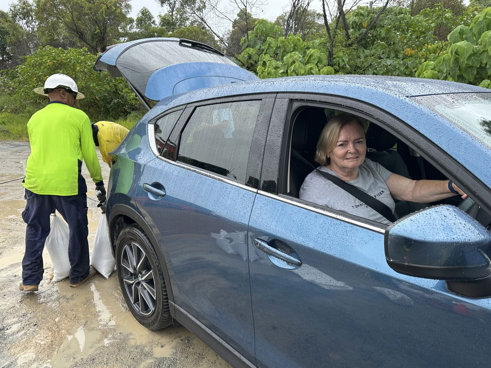 A woman in a blue car while two people put things in the boot. 