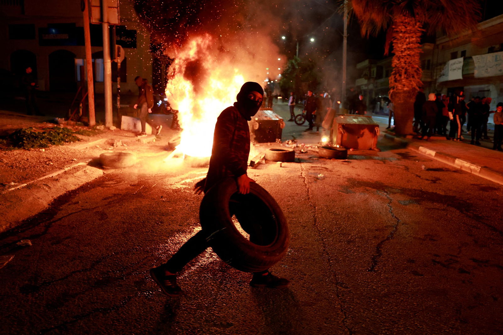 A young boy in a balaclava holding a tire walks in front of a large fire in a road.