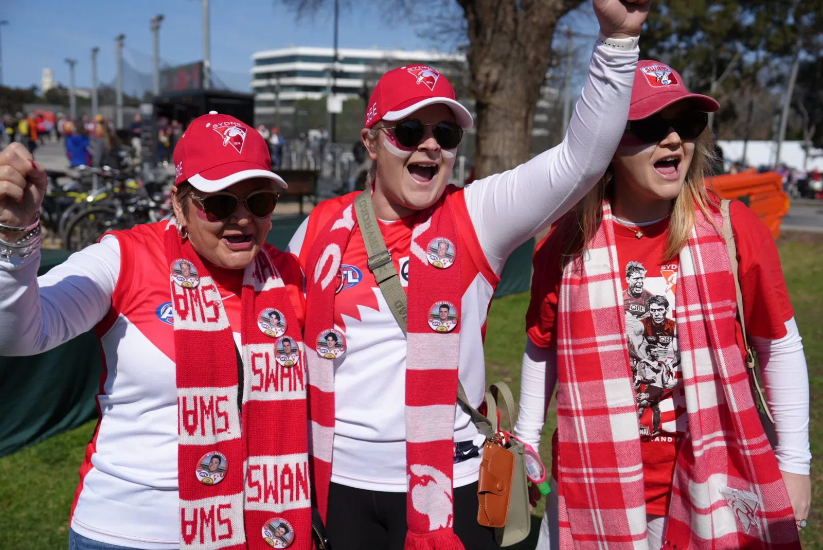 Three women decked in Sydney Swans colours raise their arms and cheer.