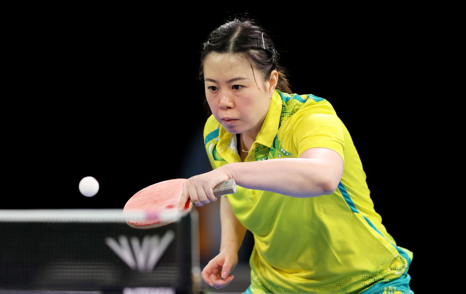 A woman with a yellow shirt and tied up hair holds a table tennis bat as a table tennis ball comes over a table tennis net. It is a black background.