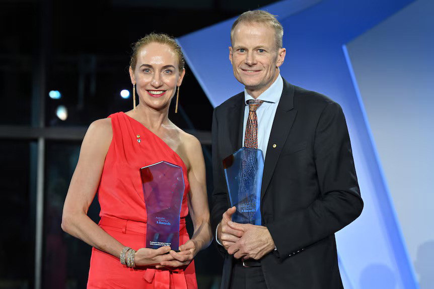 Georgina con un vestido rojo brillante sosteniendo un premio y Richard a su derecha con un traje también sosteniendo un premio. Ambos sonríen