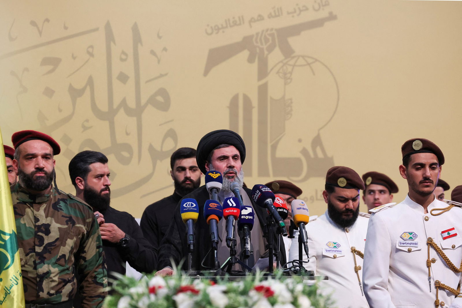 A man with a grey beard and black turban speaks into microphones while surrounded by security guards.