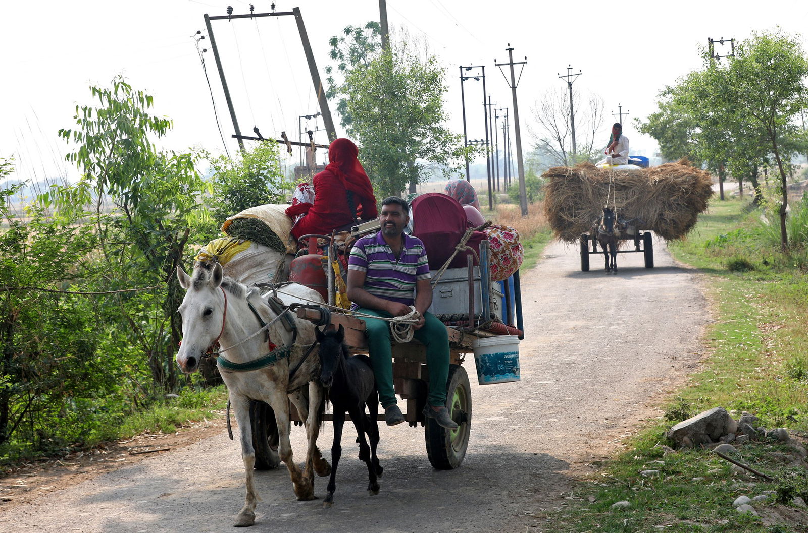 A man guides a donkey drawing a card carrying two people and goods down a dirt road with another donkey dragging hay following along in the background