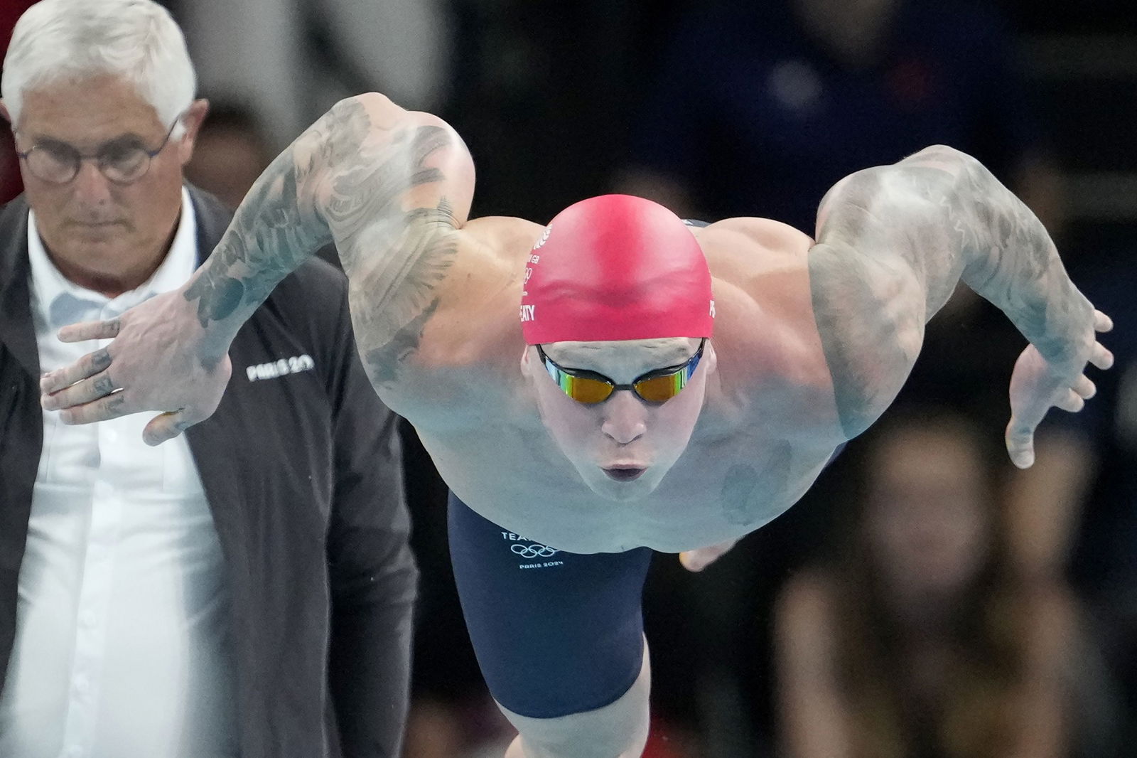 A swimmer in a red cap and gold gogggles leaps dives front on towards the camera.