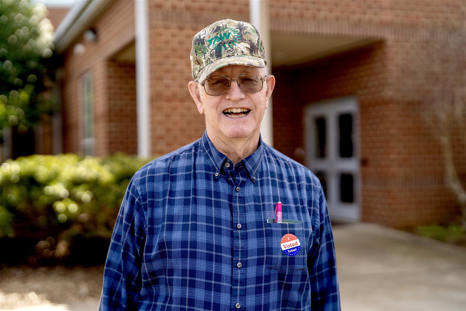 Charles Jones stands outside a brick building, wearing a cap and an 'I voted today' sticker on a blue checked shirt.