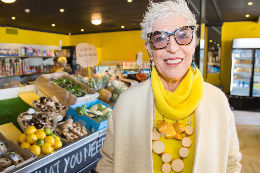 A woman in a beige cardigan, yellow top, scarf, beaded necklace and dark-rimmed sunglasses smiles in the supermarket aisle.