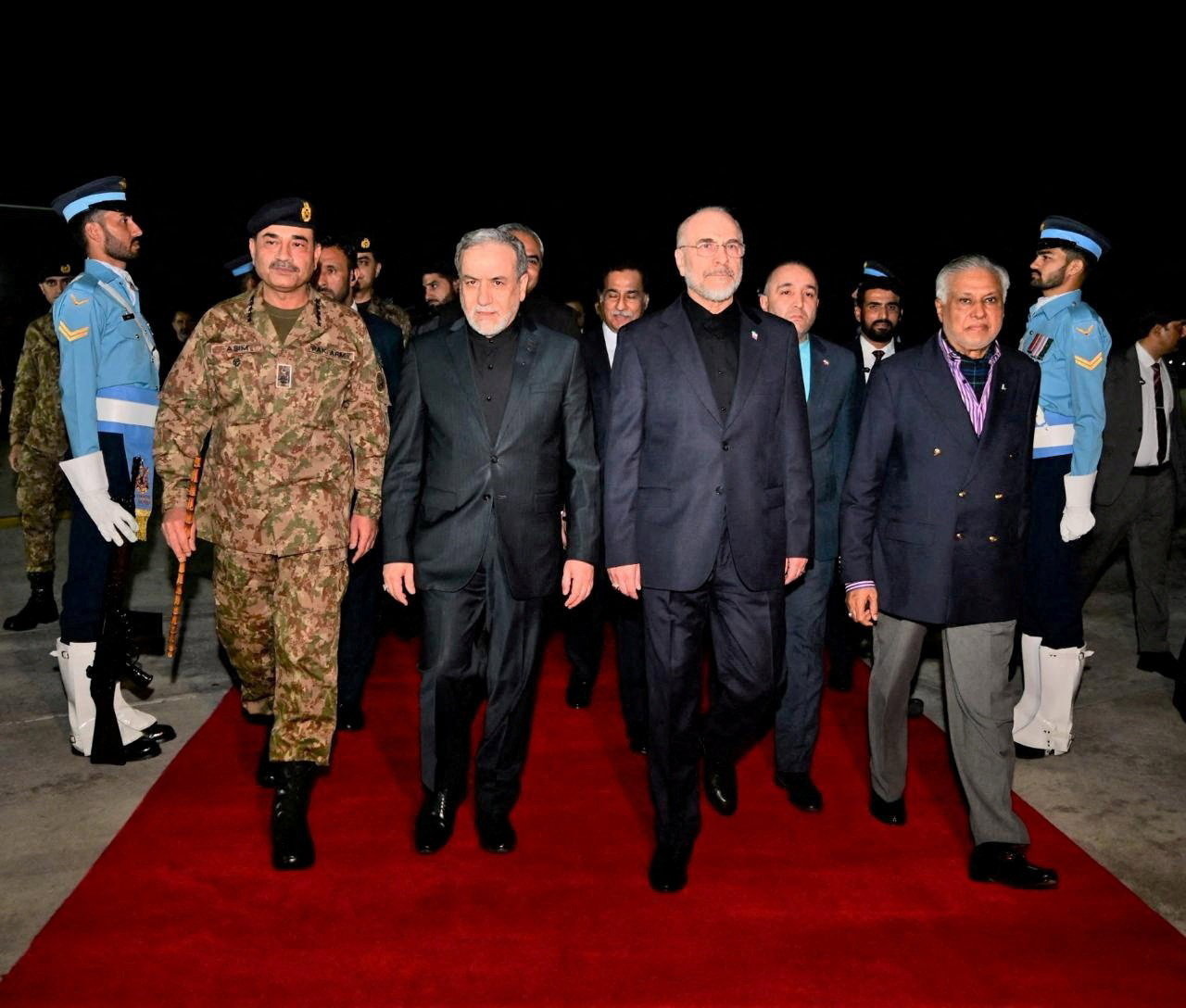 Men in suits walk down a red carpet at night