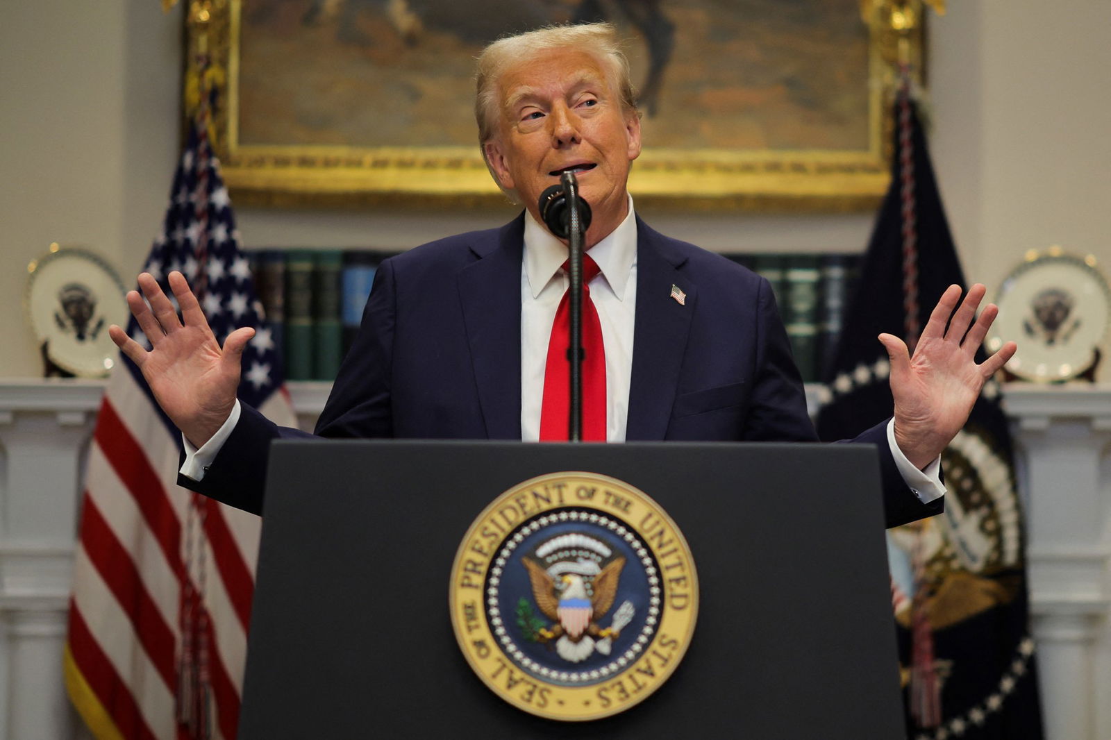 Donald trump wearing a blue suit, white shirt and red tie with hands raised standing behind a US Presidential lectern and microphone