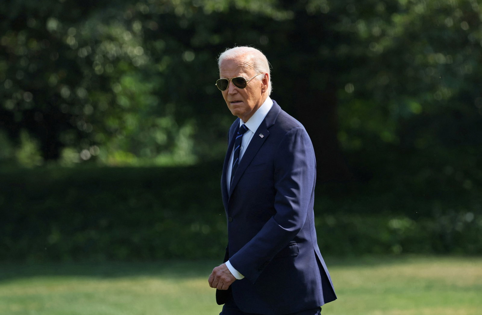 US president Joe Biden walks across the South Lawn of the White House, wearing a suit and aviator sunglasses