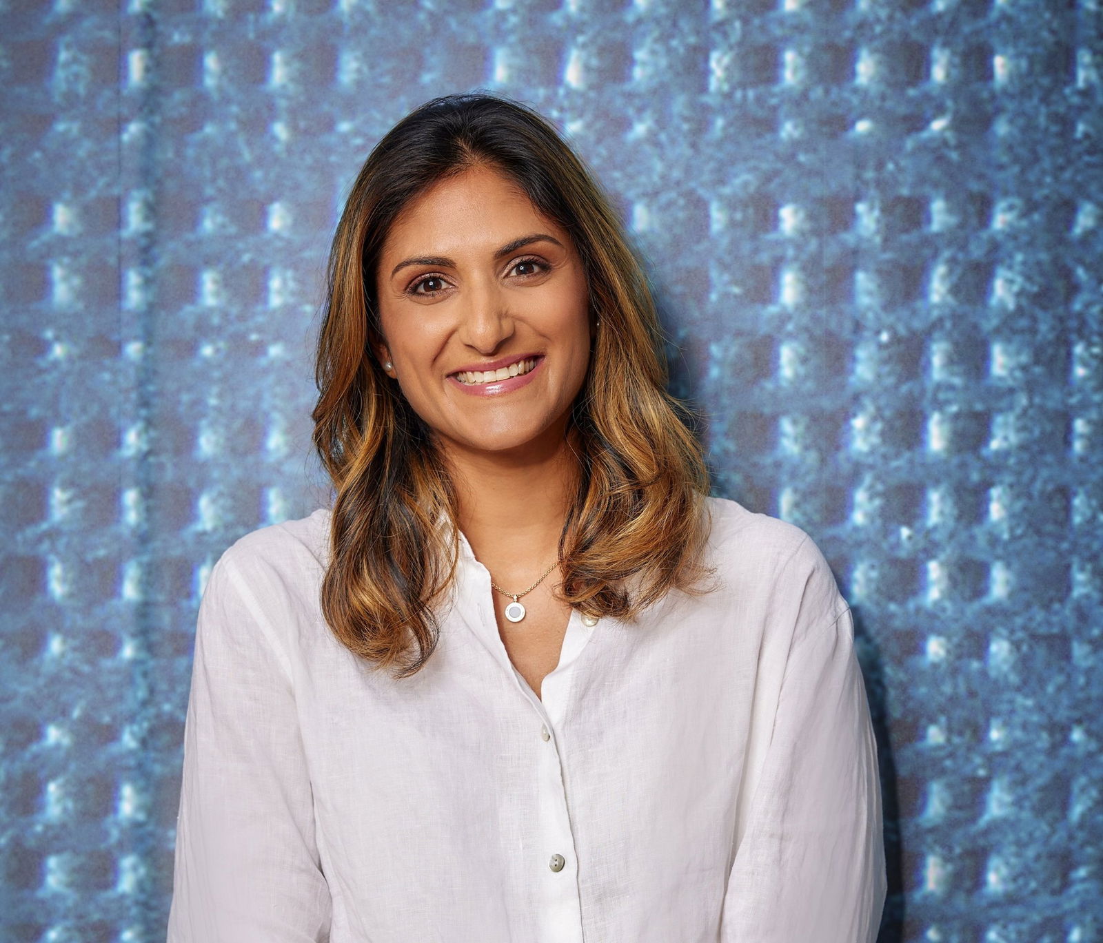 A woman in a cream button up shirt stands in front of a blue backdrop