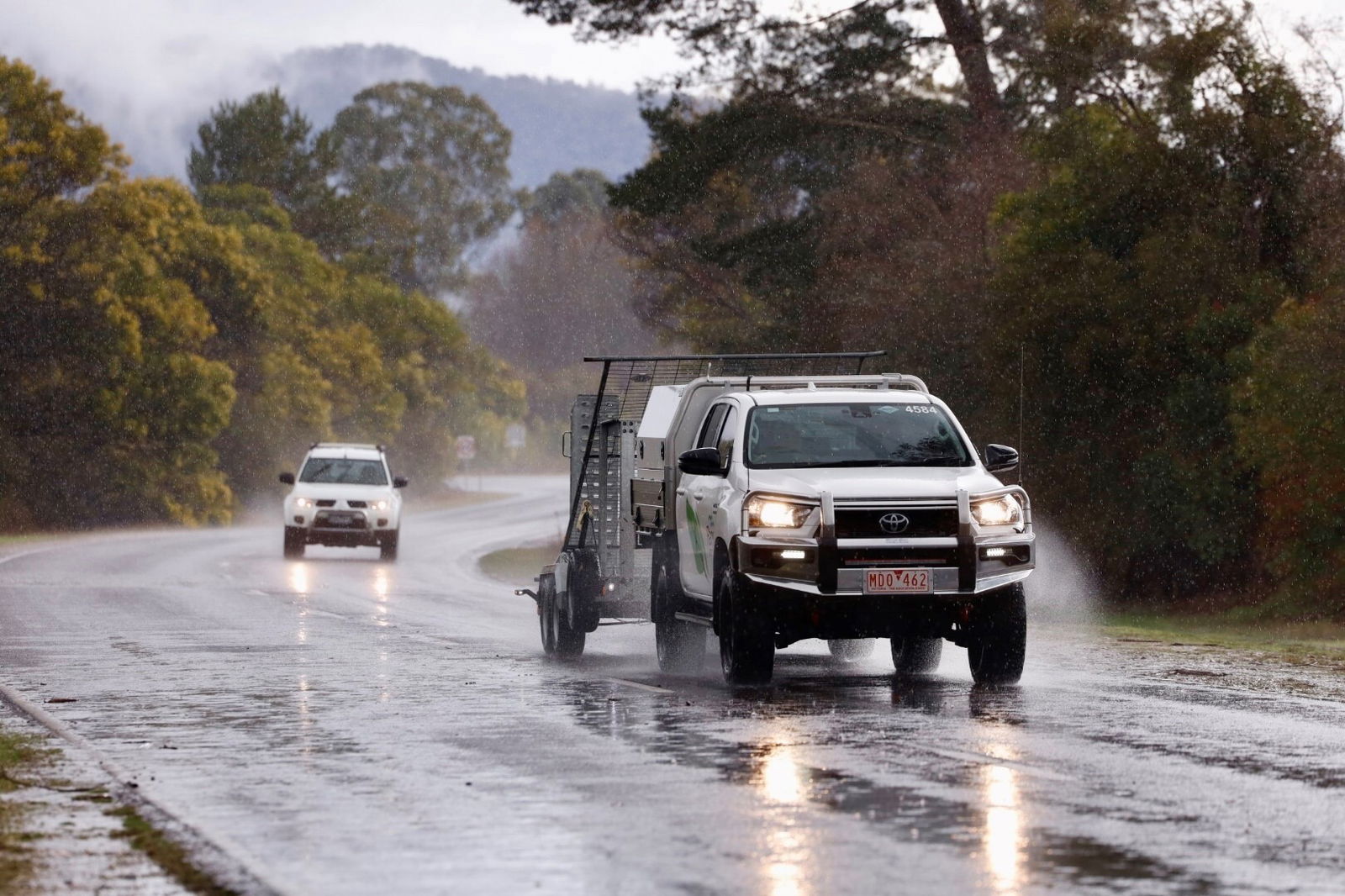 Rain falls as two cars drive along a twisted country road lined with trees.