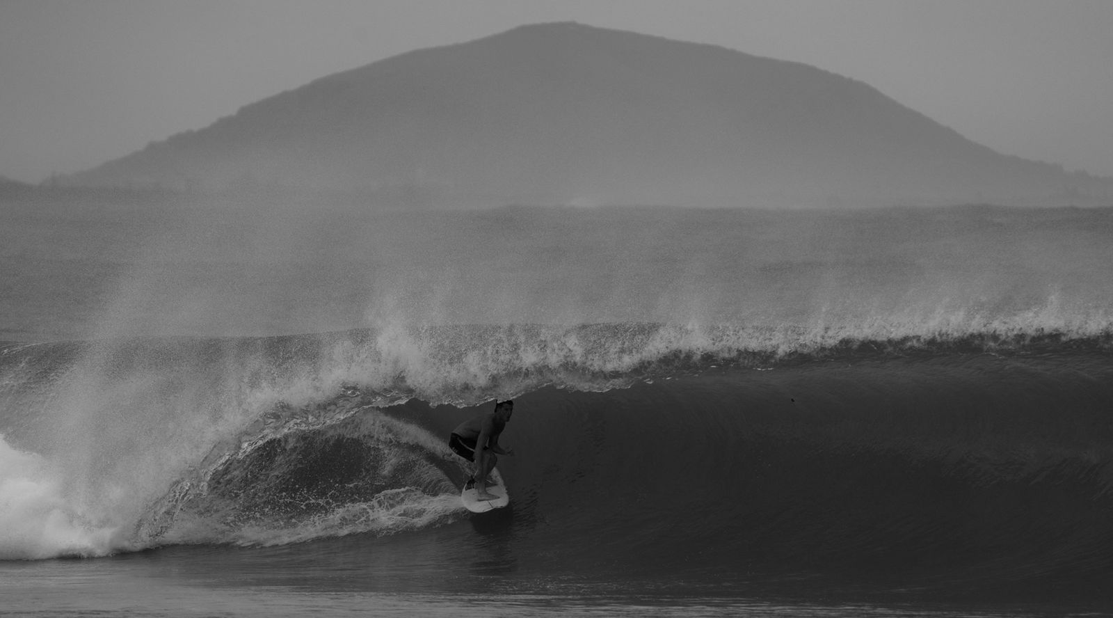 A black and white photo of a man surfing, the wave breaking over his head to create a tube of water.