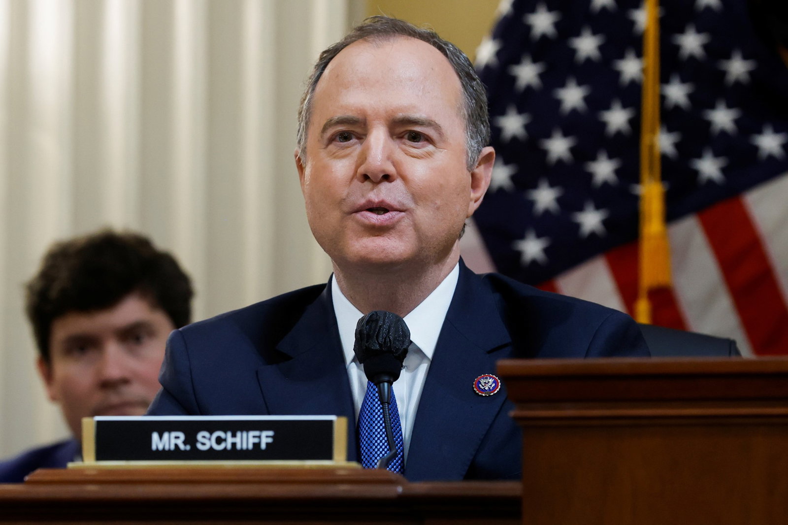 Adam Schiff speaks into a microphone while sitting at a desk behind a nameplate
