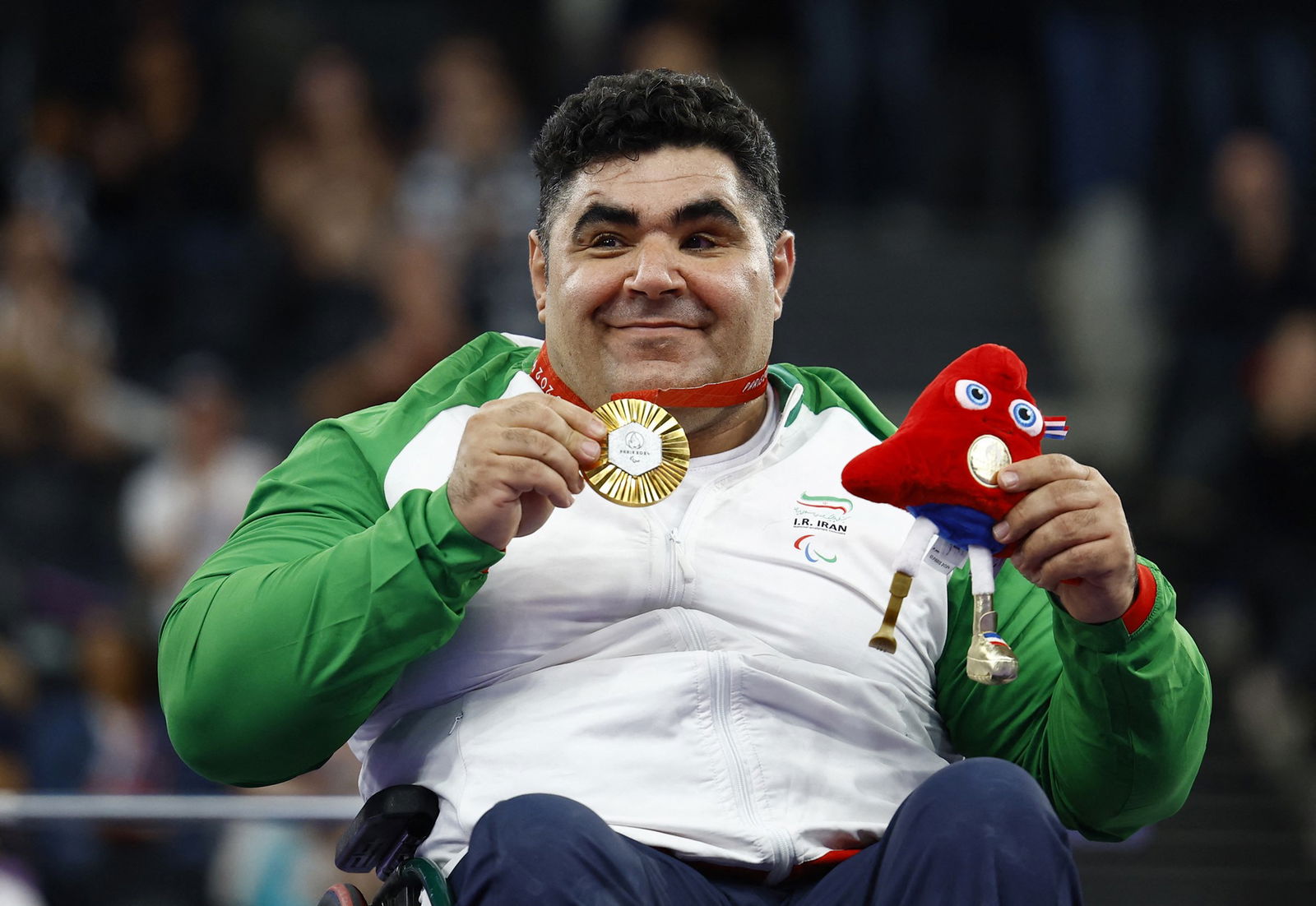 Iranian powerlifter Aliakbar Gharibshani sits in his wheelchair and smiles while holding a gold medal and a small plush toy.
