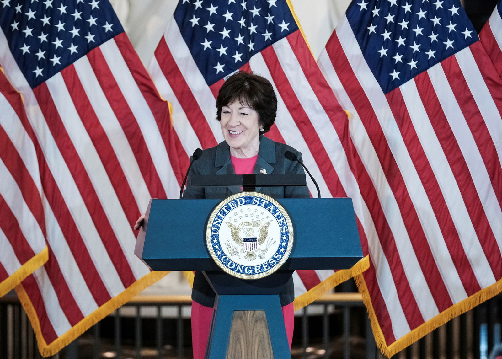 Susan Collins speaking at a lecturn in front of American flags. 