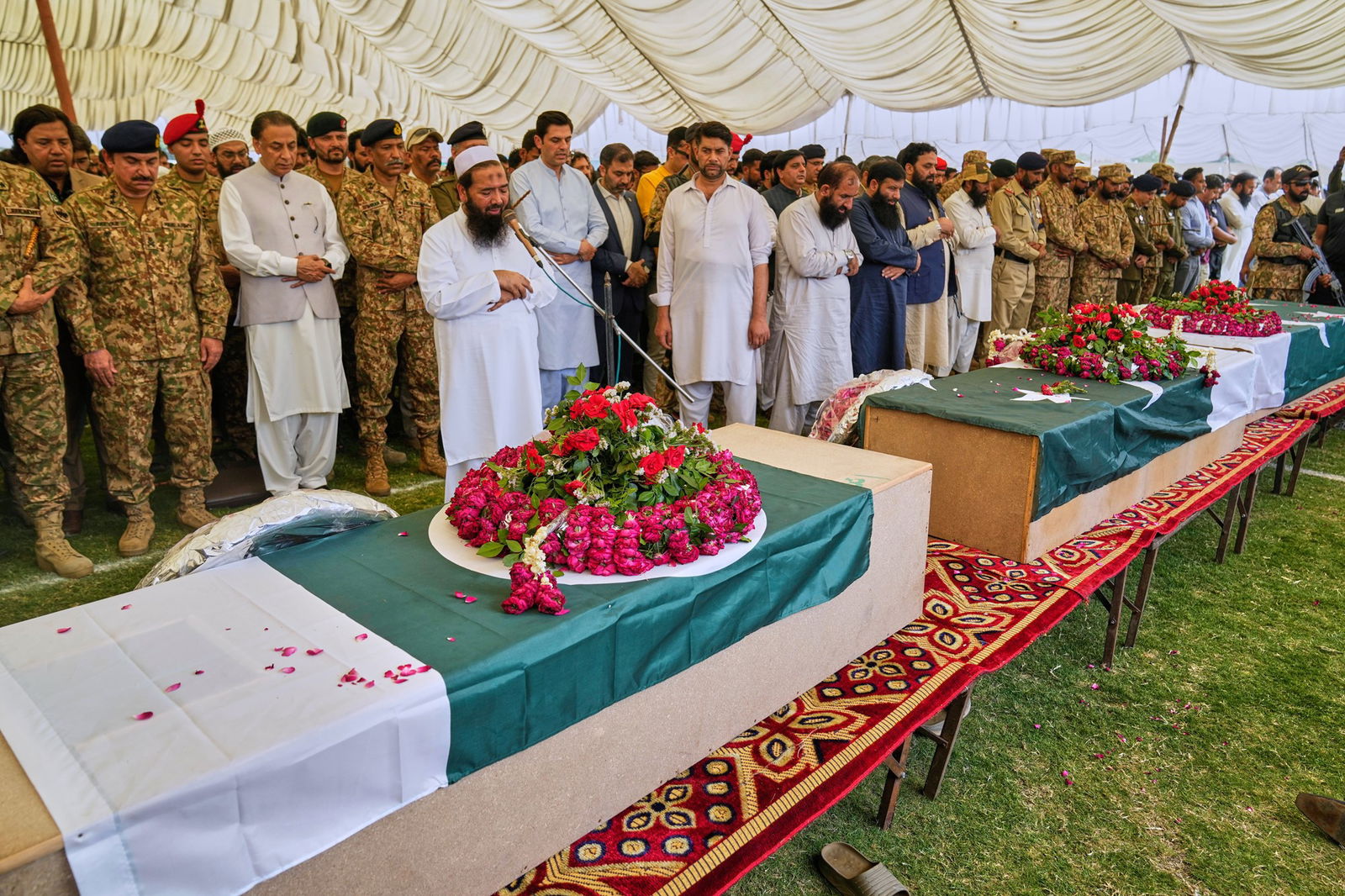 Coffins sit on table drapped in Pakistani flags in front of a religious leader who is speaking into a microphone with people standing in honour behind him
