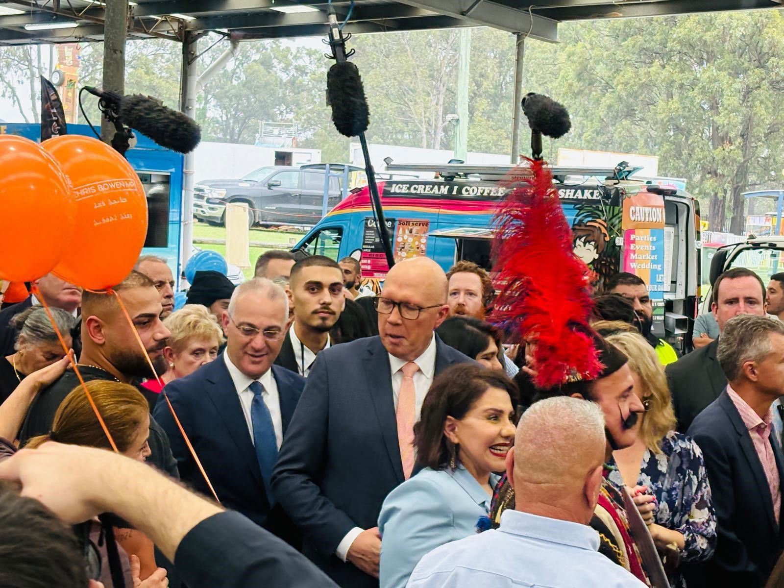 Peter Dutton is surrounded by a crowd, some carrying orange balloons and others boom microphones.